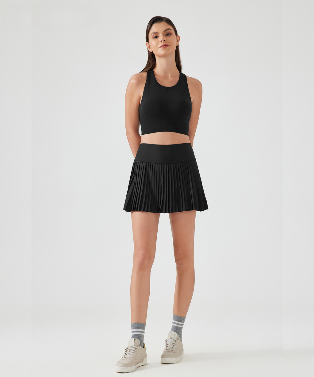 Woman wearing a black athletic tank top and a black high-rise pleated pickleball skirt on a white background